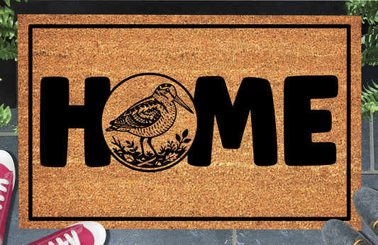 American Woodcock coir doormat in entryway — Bogsucker full-body illustration in circular botanical frame replaces the letter O in HOME on natural coir