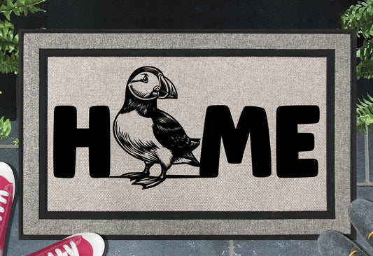 Atlantic Puffin all-weather doormat in a doorway — full-body standing Puffin illustration replacing the letter O in "HOME", UV-cured inks on woven polyester, front door entryway