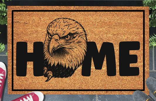American Bald Eagle HOME doormat in doorway lifestyle shot — natural tan coir mat, large angled Bald Eagle head and talon illustration replaces the letter O in HOME, 18x30 size shown
