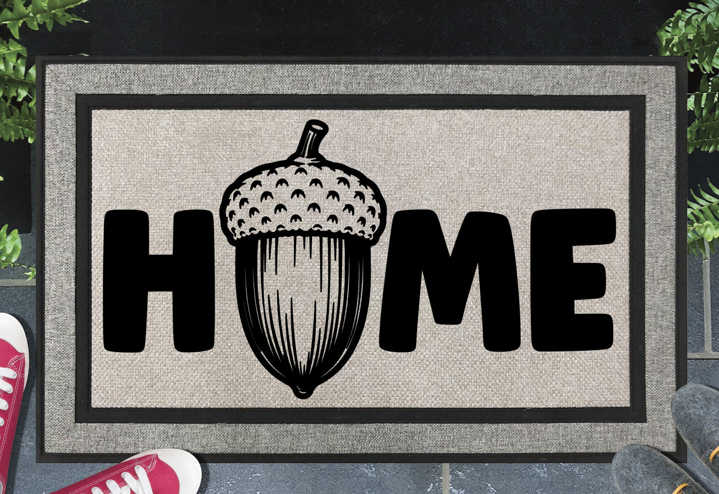 English Oak Pedunculate Oak acorn all-weather doormat with bold black HOME lettering and large detailed acorn illustration replacing the letter O, on a grey woven polyester linen-look surface with black border, styled on a slate doorstep. 