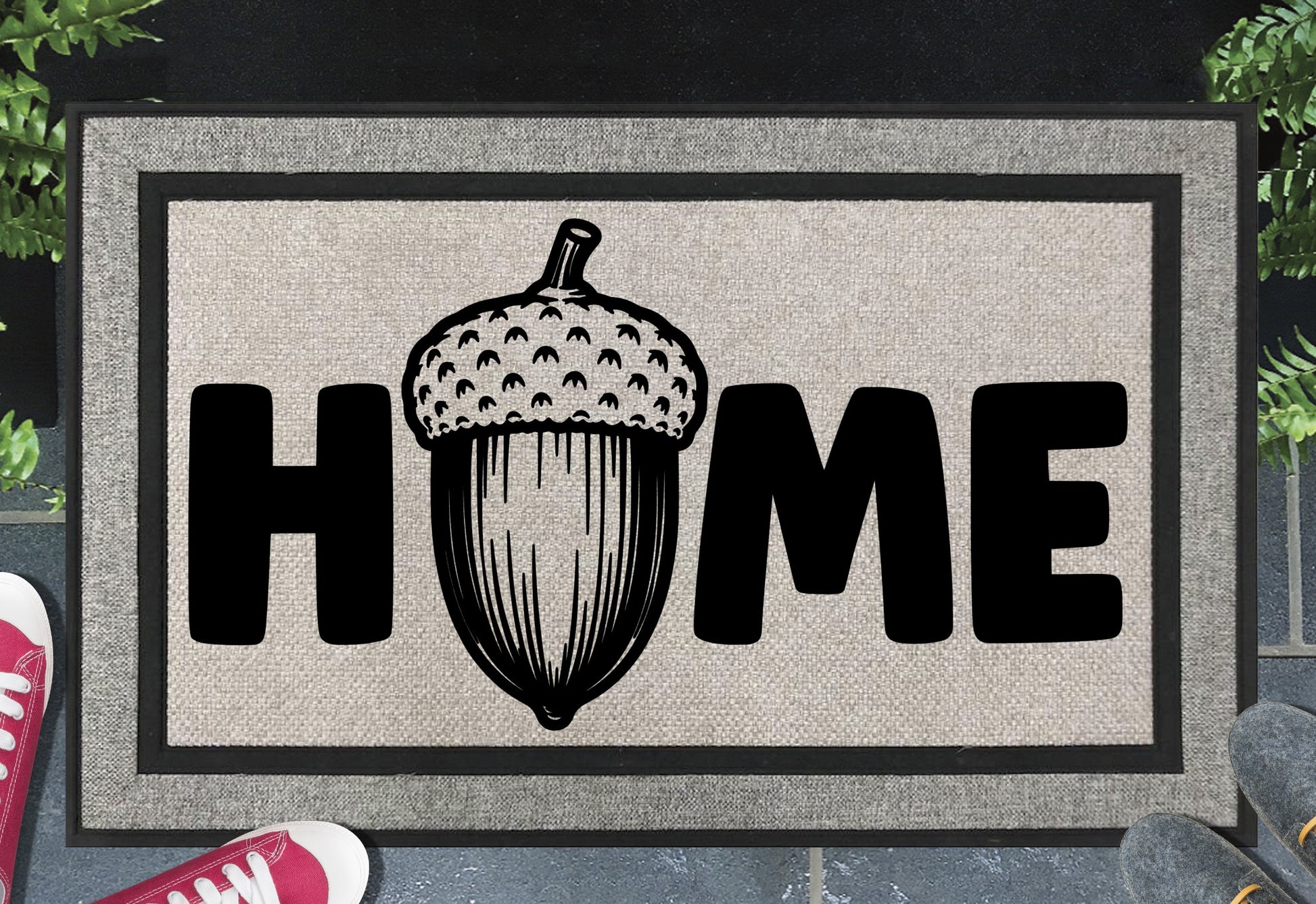 English Oak Pedunculate Oak acorn all-weather doormat with bold black HOME lettering and large detailed acorn illustration replacing the letter O, on a grey woven polyester linen-look surface with black border, styled on a slate doorstep. 