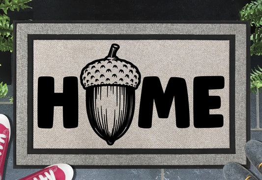 English Oak Pedunculate Oak acorn all-weather doormat with bold black HOME lettering and large detailed acorn illustration replacing the letter O, on a grey woven polyester linen-look surface with black border, styled on a slate doorstep. 