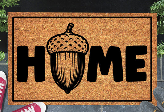 English Oak Pedunculate Oak acorn coir doormat with bold black HOME lettering and detailed acorn illustration replacing the letter O, on a natural golden coir surface with black border, styled on a slate doorstep.