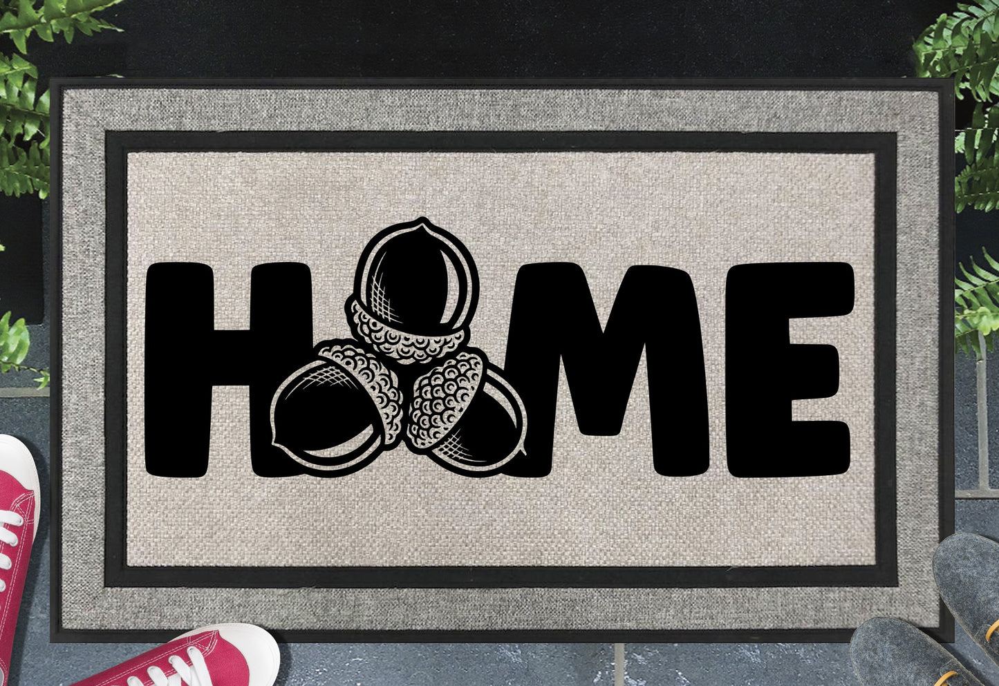 Sessile Oak Welsh Oak acorn all-weather doormat with bold black HOME lettering and cluster of three acorn illustrations replacing the letter O, on a grey woven polyester linen-look surface with black border, styled on a slate doorstep. 