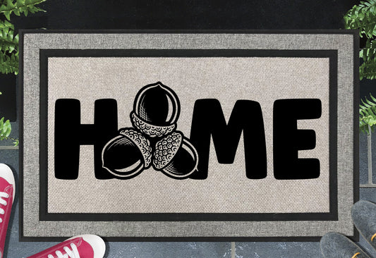 Sessile Oak Welsh Oak acorn all-weather doormat with bold black HOME lettering and cluster of three acorn illustrations replacing the letter O, on a grey woven polyester linen-look surface with black border, styled on a slate doorstep. 