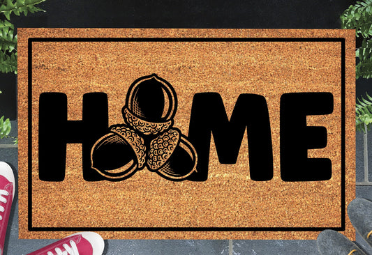Sessile Oak Welsh Oak acorn coir doormat with bold black HOME lettering and cluster of three acorn illustrations replacing the letter O, on a natural golden coir surface with black border, styled on a slate doorstep.
