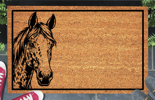 Appaloosa Horse peeking from the left edge of a natural coir doormat, with a spotted face, sweeping mane and black border, shown at an entryway.