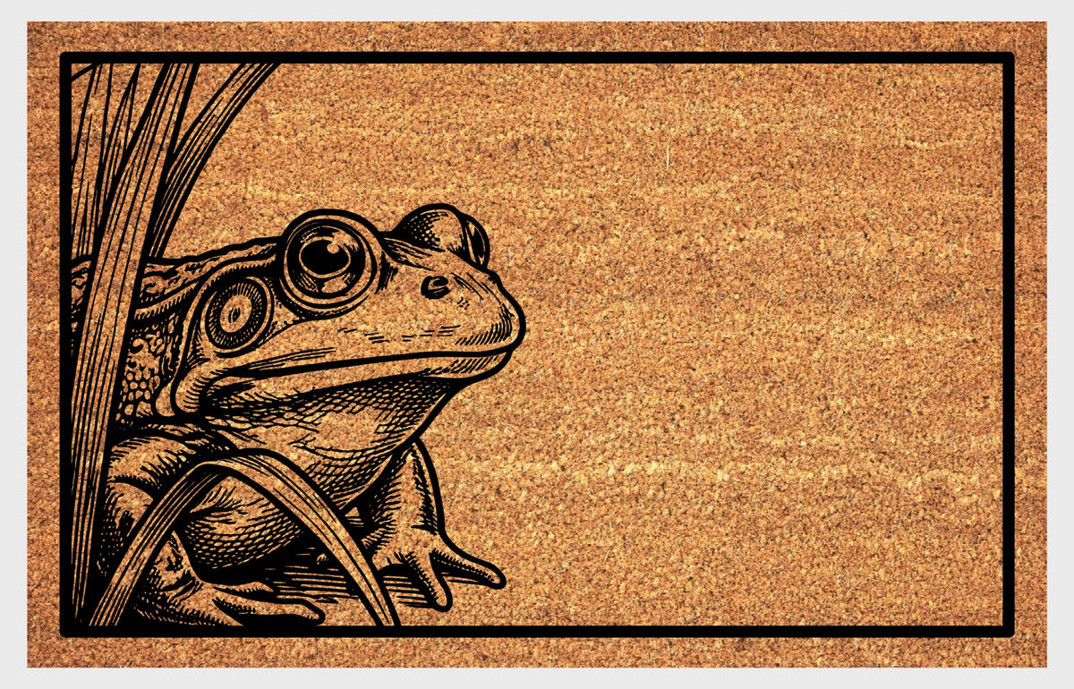 Close-up of a natural coir doormat featuring a wide-eyed Bullfrog peeking from the left edge among reeds.