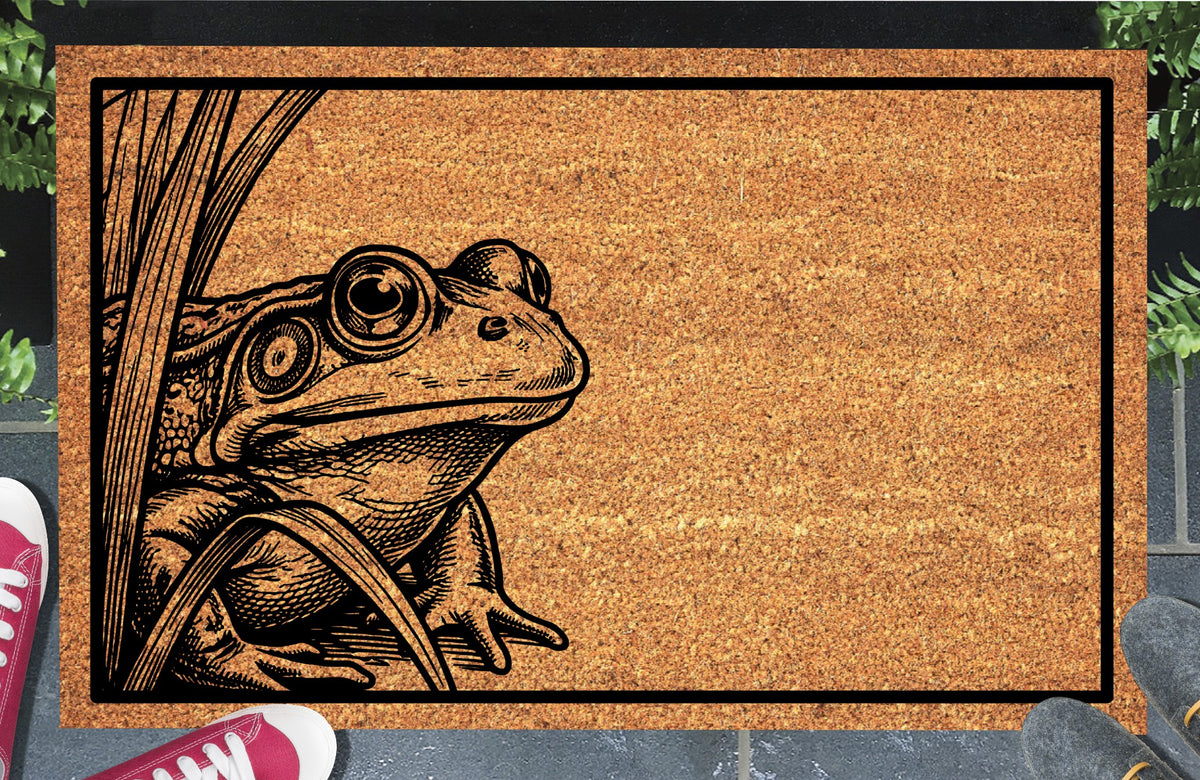 Bullfrog peeking among reeds on a natural coir doormat with a black rectangular border, shown at an entryway.