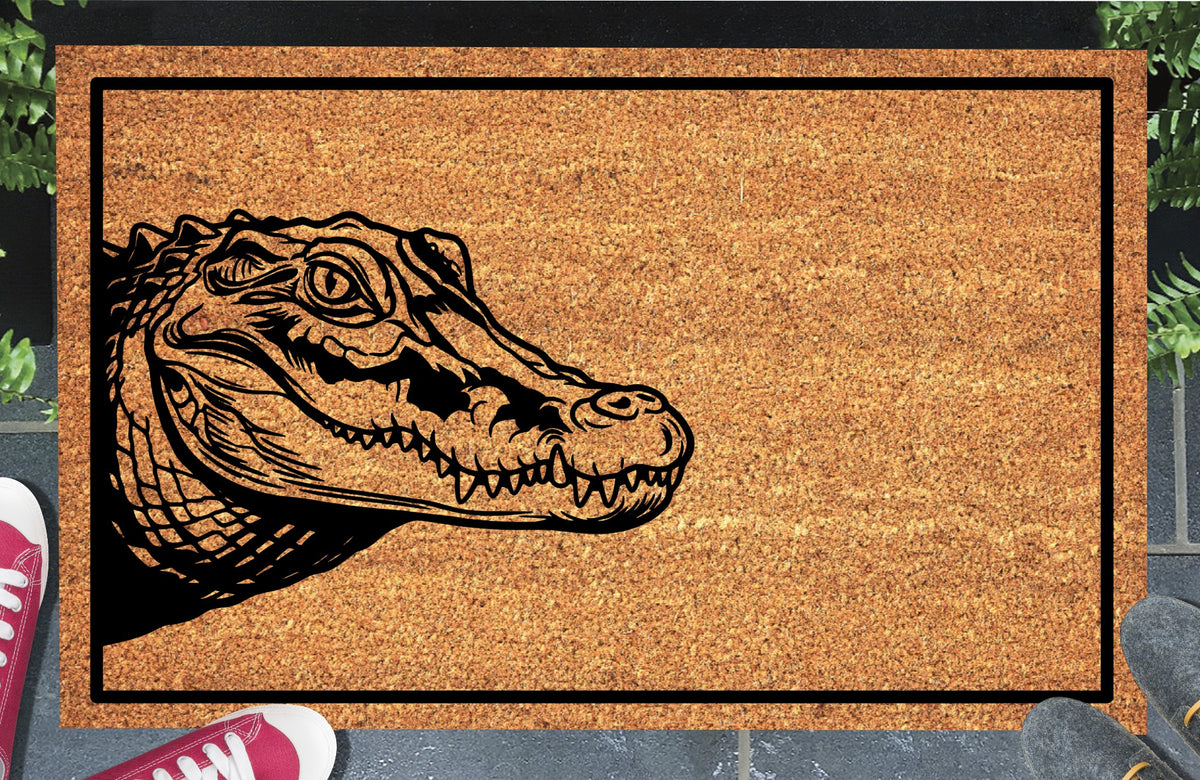 Crocodile peeking from the left edge of a natural coir doormat with closed jaws and a black border, shown at an entryway.
