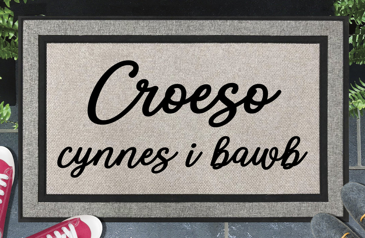 Croeso cynnes i bawb all-weather doormat — Welsh warm welcome to all in bold two-line script lettering, woven polyester, linen-look finish, non-slip backing, 18x30"