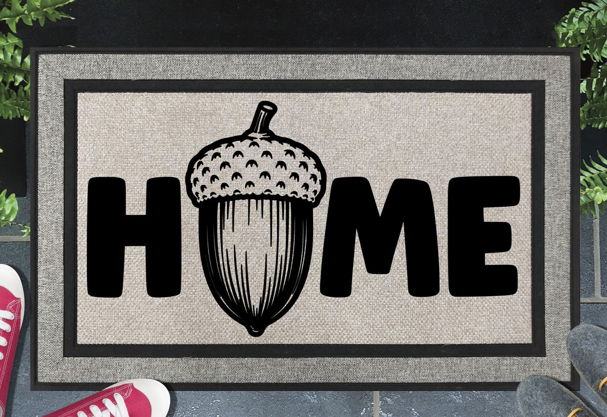 English Oak Pedunculate Oak acorn all-weather doormat with bold black HOME lettering and large detailed acorn illustration replacing the letter O, on a grey woven polyester linen-look surface with black border, styled on a slate doorstep. 