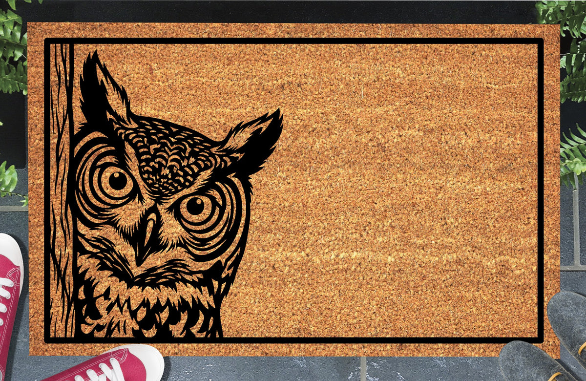 Great Horned Owl peeking coir doormat with tufted owl illustration beside a wood-grain edge and black rectangular border, styled at an entryway.