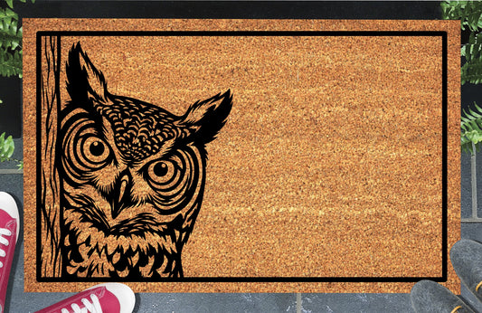 Great Horned Owl peeking coir doormat with tufted owl illustration beside a wood-grain edge and black rectangular border, styled at an entryway.