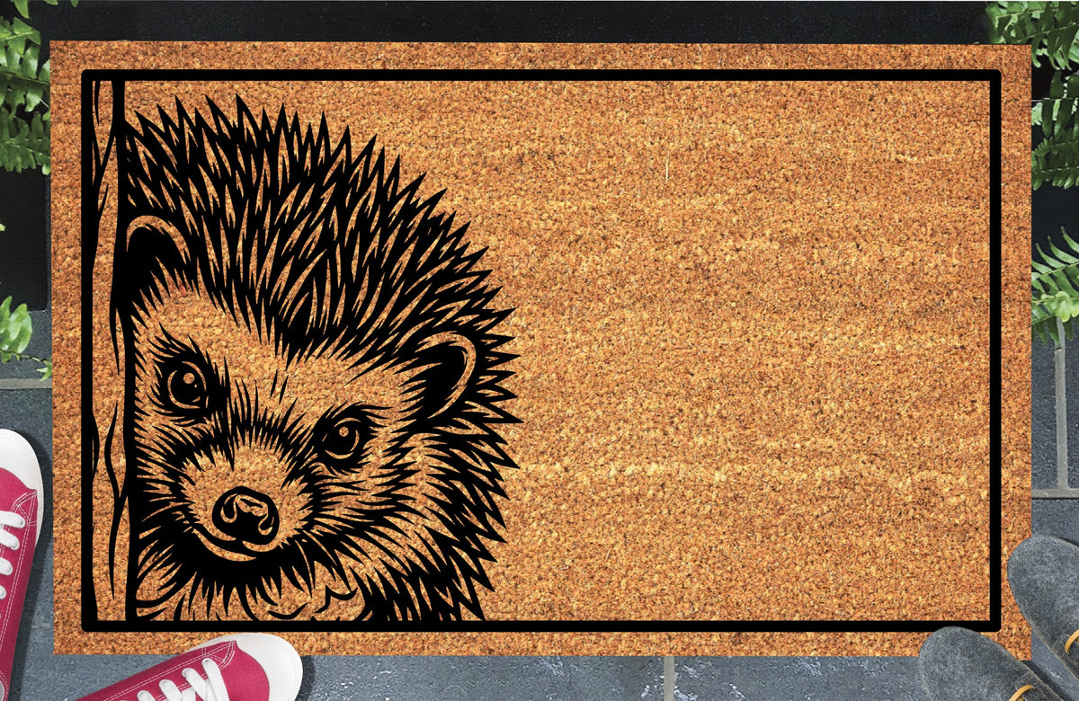 Hedgehog peeking coir doormat with a spiny hedgehog beside a wood-grain edge and black rectangular border, styled at an entryway.