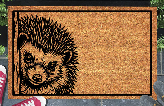Hedgehog peeking coir doormat with a spiny hedgehog beside a wood-grain edge and black rectangular border, styled at an entryway.
