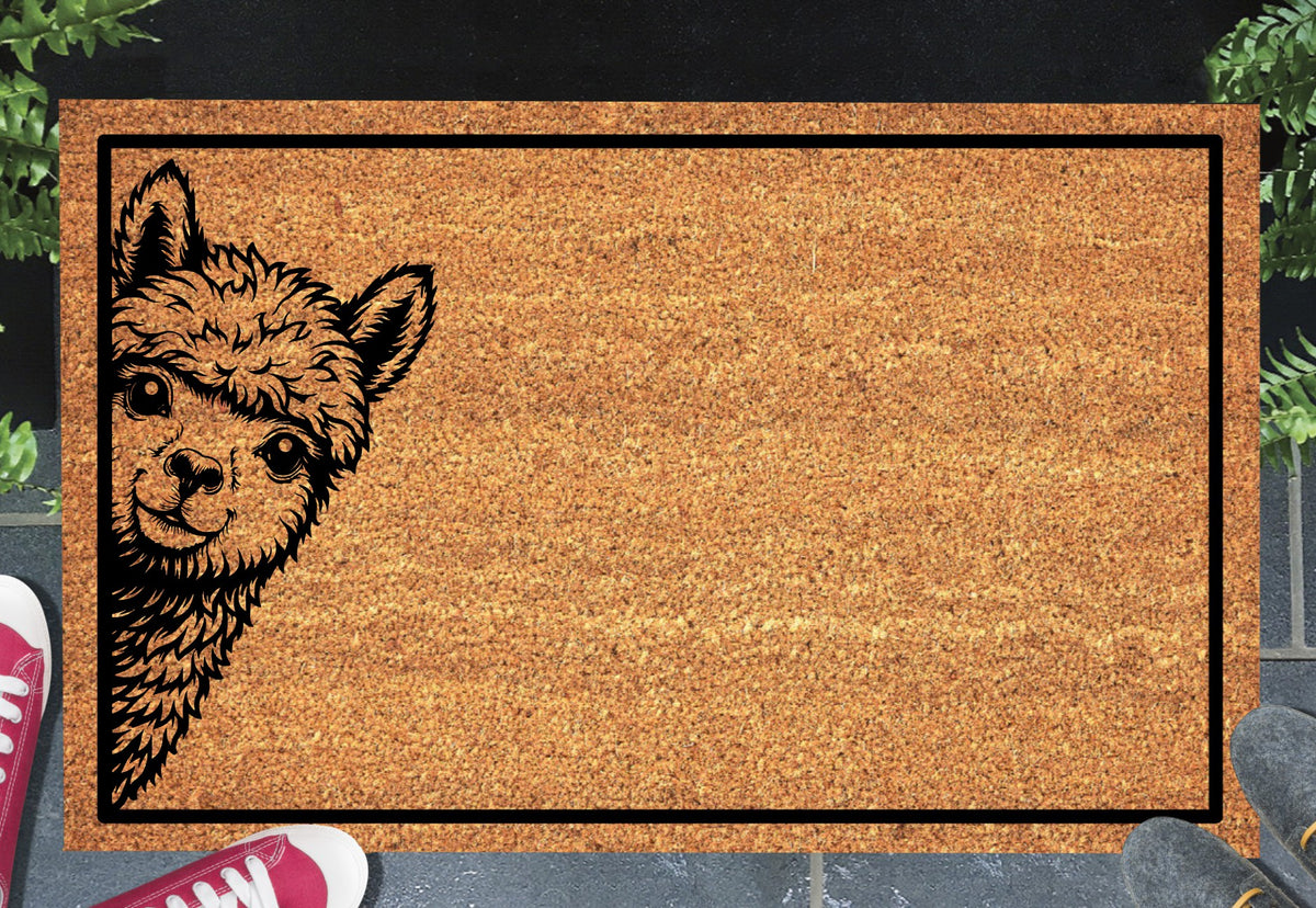 Huacaya Alpaca peeking coir doormat with a smiling Alpaca peeking from the left edge inside a black rectangular border, styled at an entryway.
