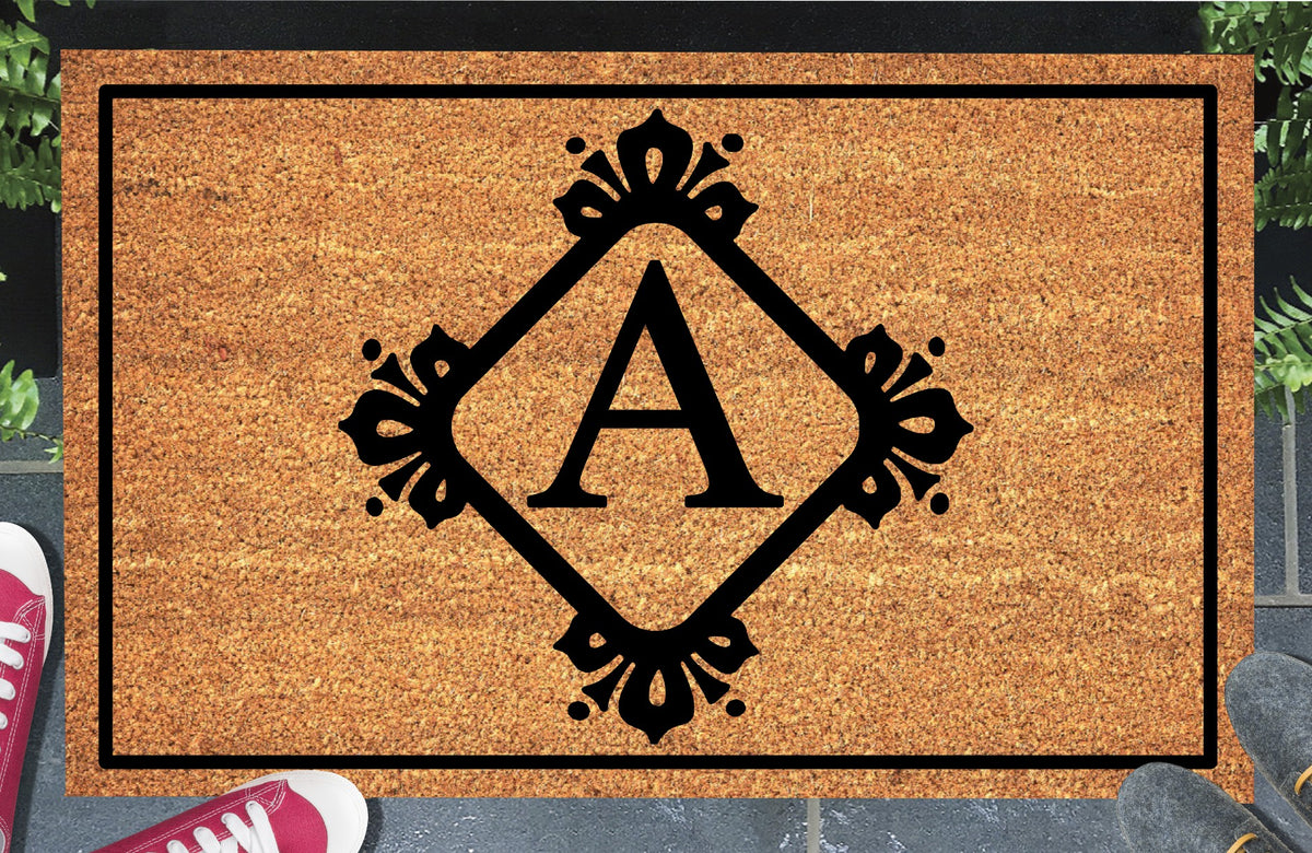 Monogram Letter A doormat in natural coir, showing the ornate serif letter A in a diamond frame with fleur-de-lis style flourishes and dot accents, with a black border, displayed on a doorstep. 