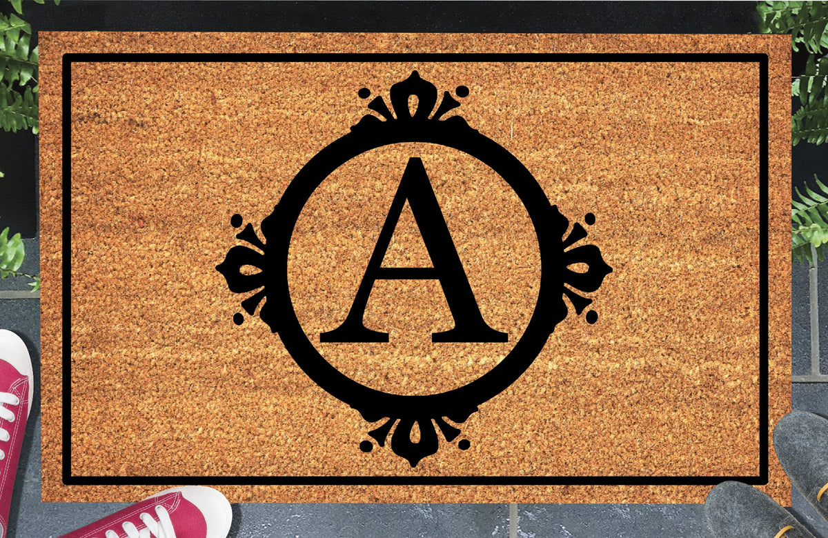 Monogram Letter A doormat in natural coir, featuring a serif A in a circular frame with fleur-de-lis style flourishes, dot accents and a black rectangular border, displayed on a doorstep.