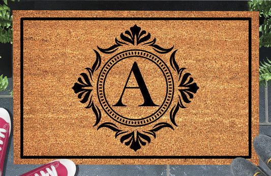 Monogram Letter A doormat in natural coir, showing the ornate serif letter A in a dotted circular frame with fleur-de-lis style flourishes and a black border, displayed on a doorstep. 