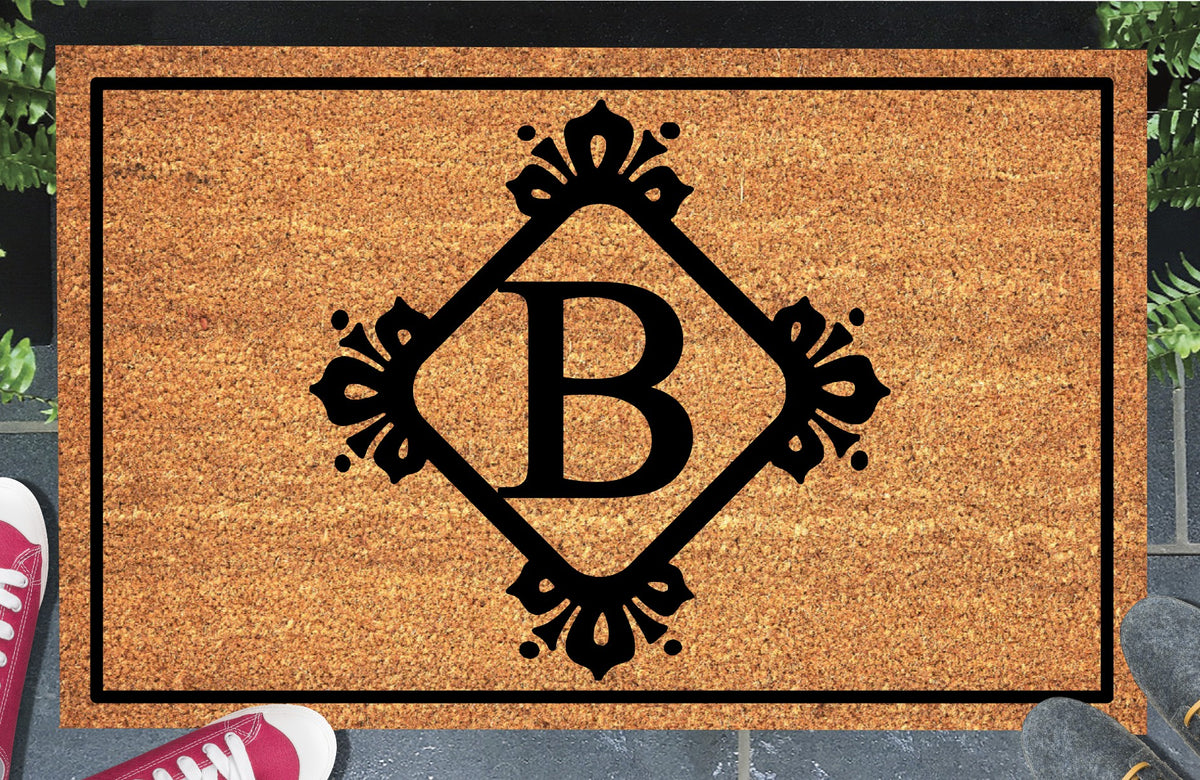 Monogram Letter B doormat in natural coir, showing the ornate serif letter B in a diamond frame with fleur-de-lis style flourishes and dot accents, with a black border, displayed on a doorstep.  
