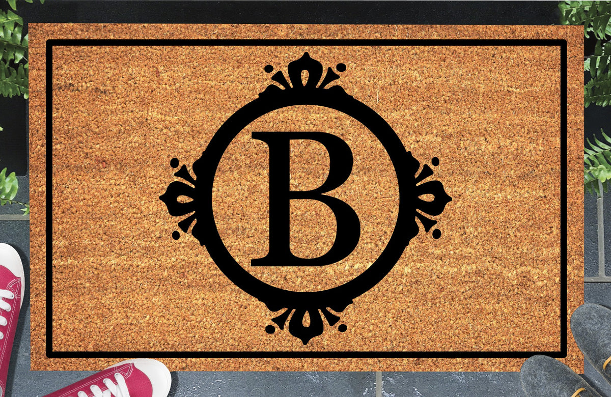 Monogram Letter B doormat in natural coir, featuring a serif B in a circular frame with fleur-de-lis style flourishes, dot accents and a black rectangular border, displayed on a doorstep.