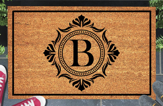 Monogram Letter B doormat in natural coir, showing the ornate serif letter B in a dotted circular frame with fleur-de-lis style flourishes and a black border, displayed on a doorstep. 