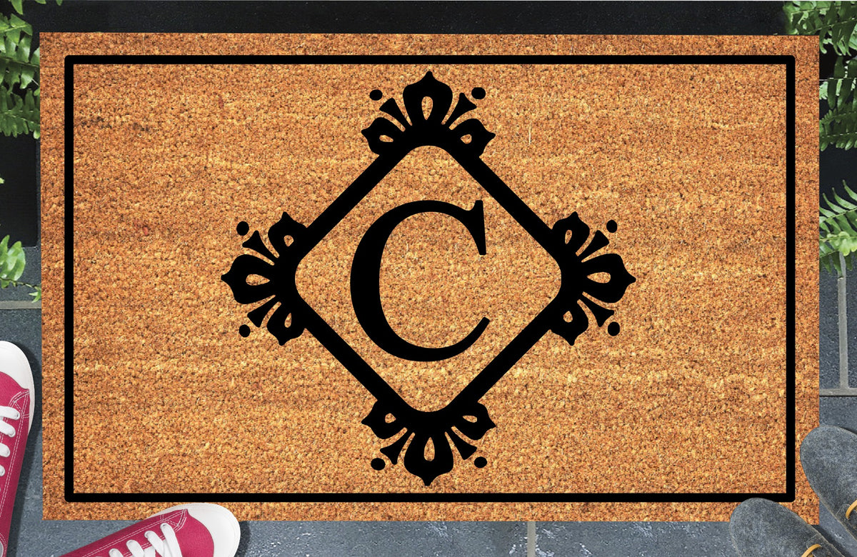 Monogram Letter C doormat in natural coir, showing the ornate serif letter C in a diamond frame with fleur-de-lis style flourishes and dot accents, with a black border, displayed on a doorstep. 