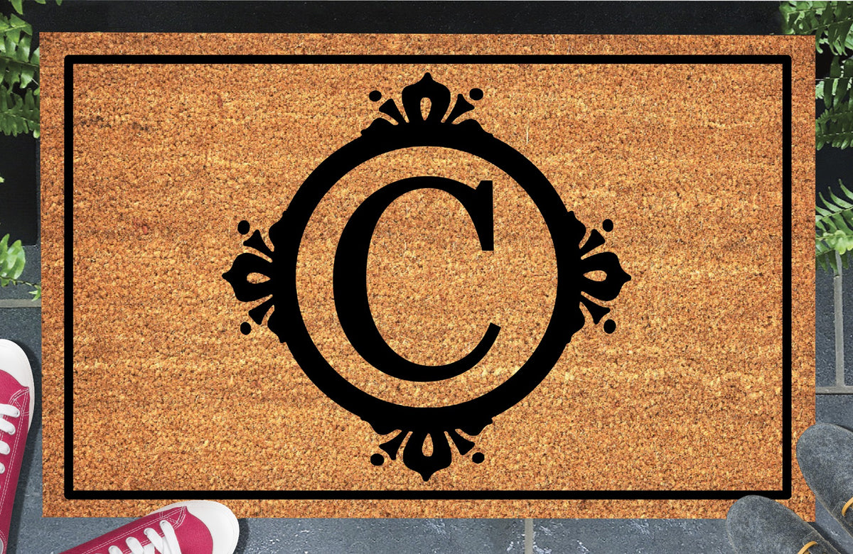 Monogram Letter C doormat in natural coir, featuring a serif C in a circular frame with fleur-de-lis style flourishes, dot accents and a black rectangular border, displayed on a doorstep.