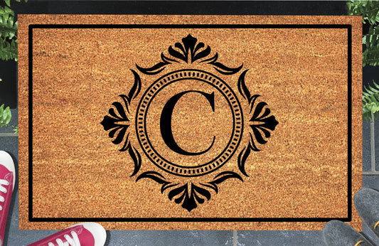 Monogram Letter C doormat in natural coir, showing the ornate serif letter C in a dotted circular frame with fleur-de-lis style flourishes and a black border, displayed on a doorstep. 