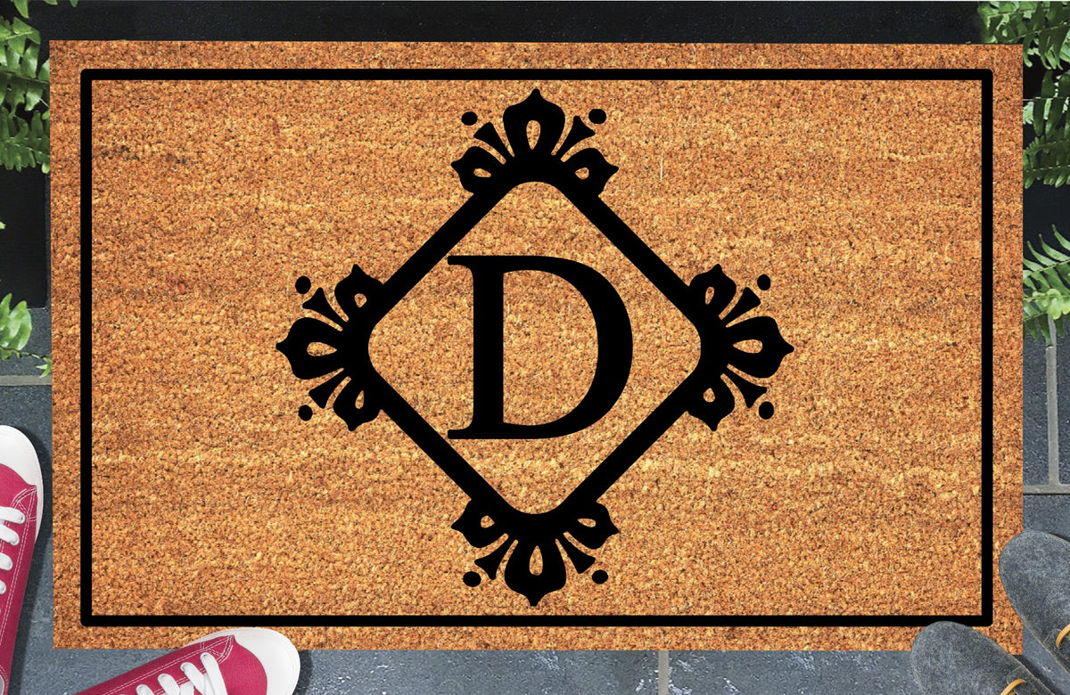 Monogram Letter D doormat in natural coir, showing the ornate serif letter D in a diamond frame with fleur-de-lis style flourishes and dot accents, with a black border, displayed on a doorstep.