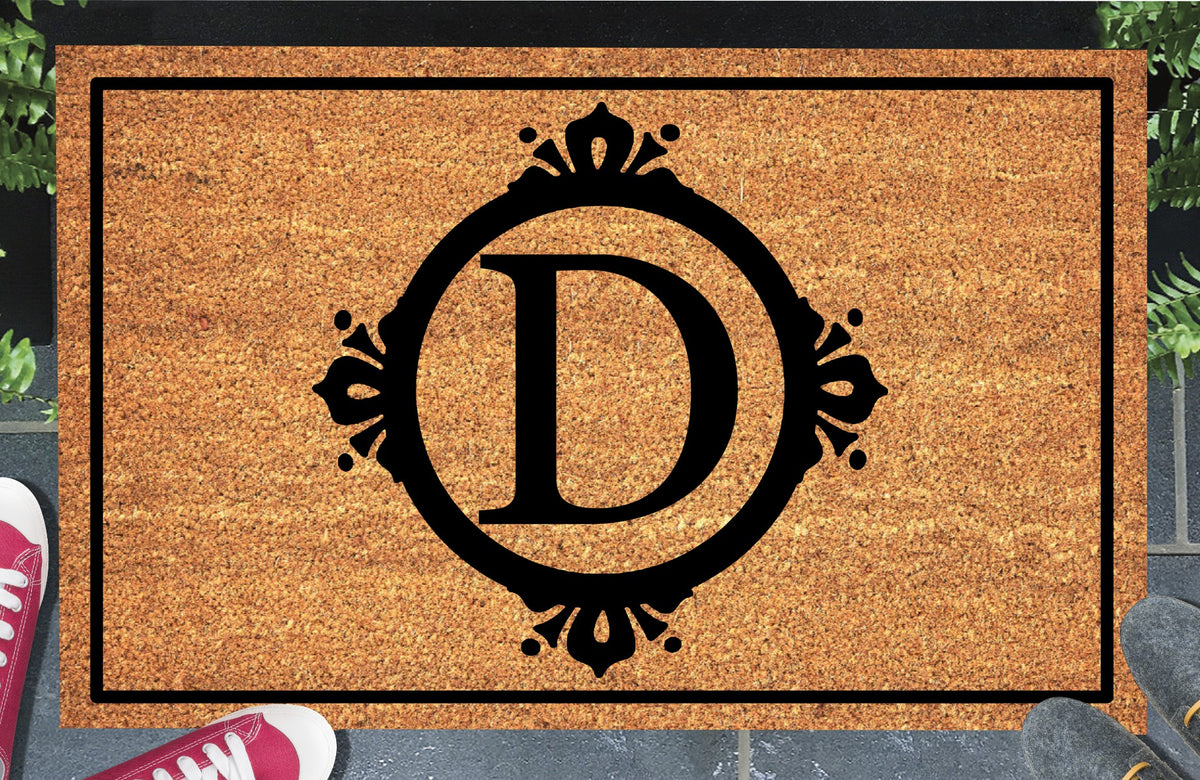 Monogram Letter D doormat in natural coir, featuring a serif D in a circular frame with fleur-de-lis style flourishes, dot accents and a black rectangular border, displayed on a doorstep.