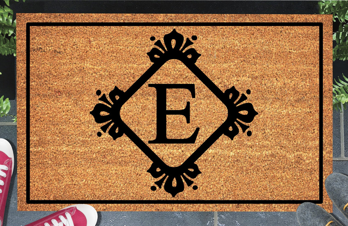 Monogram Letter E doormat in natural coir, showing the ornate serif letter E in a diamond frame with fleur-de-lis style flourishes and dot accents, with a black border, displayed on a doorstep. 