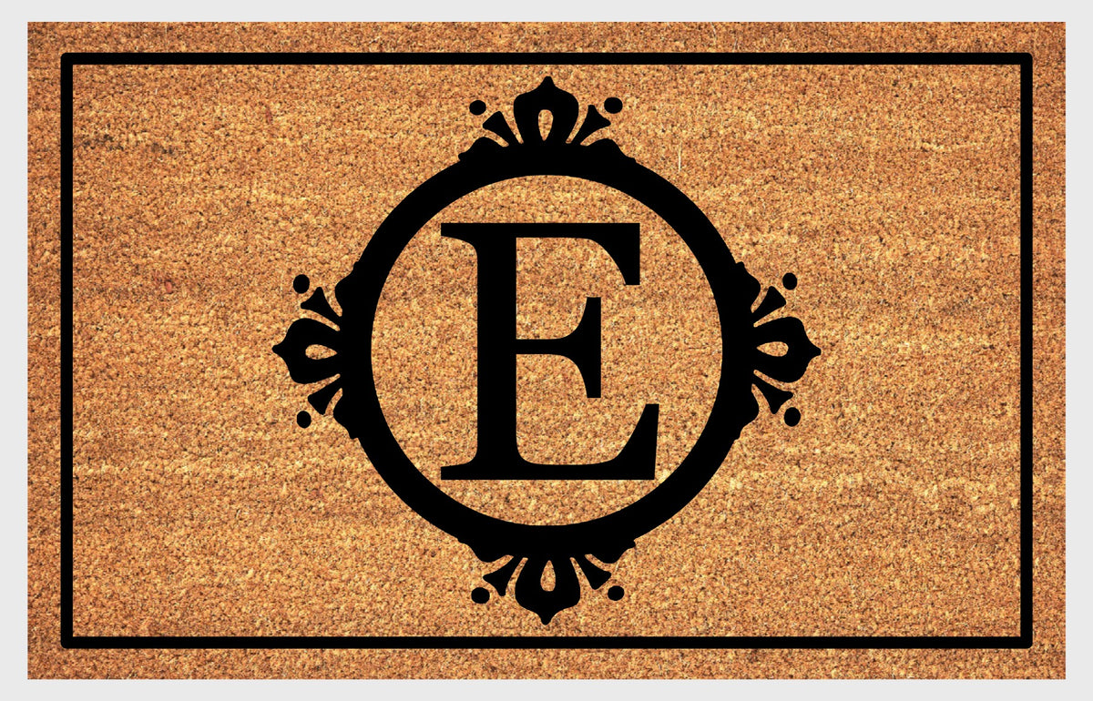 Monogram Letter E doormat in natural coir, featuring a serif E in a circular frame with fleur-de-lis style flourishes, dot accents and a black rectangular border, shown flat.