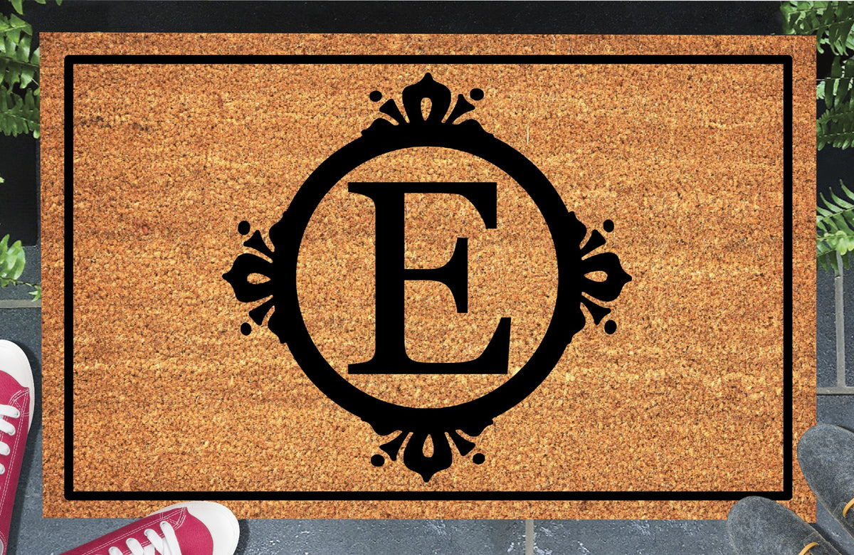Monogram Letter E doormat in natural coir, featuring a serif E in a circular frame with fleur-de-lis style flourishes, dot accents and a black rectangular border, displayed on a doorstep.
