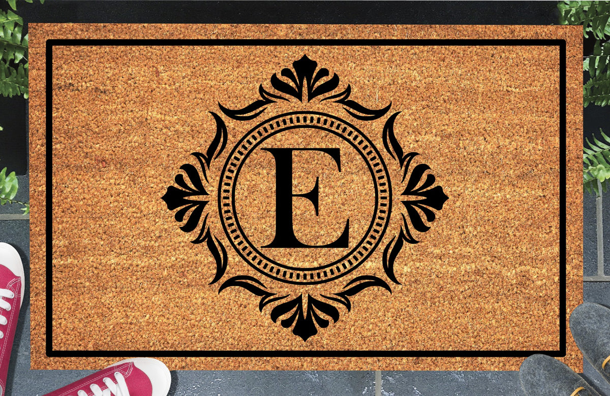 Monogram Letter E doormat in natural coir, showing the ornate serif letter E in a dotted circular frame with fleur-de-lis style flourishes and a black border, displayed on a doorstep. 