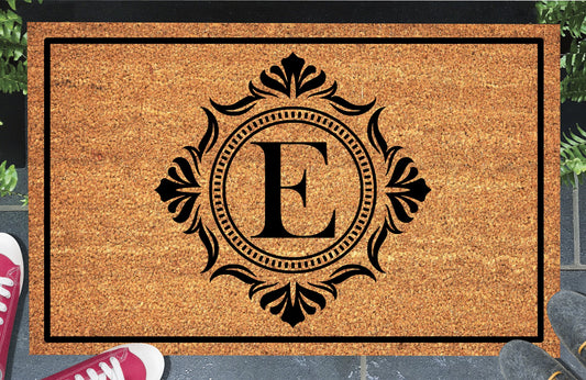 Monogram Letter E doormat in natural coir, showing the ornate serif letter E in a dotted circular frame with fleur-de-lis style flourishes and a black border, displayed on a doorstep. 
