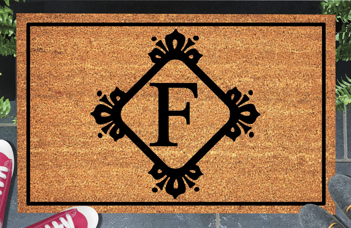 Monogram Letter F doormat in natural coir, featuring a serif F in a diamond frame with fleur-de-lis style flourishes, dot accents and a black border, displayed on a doorstep.  