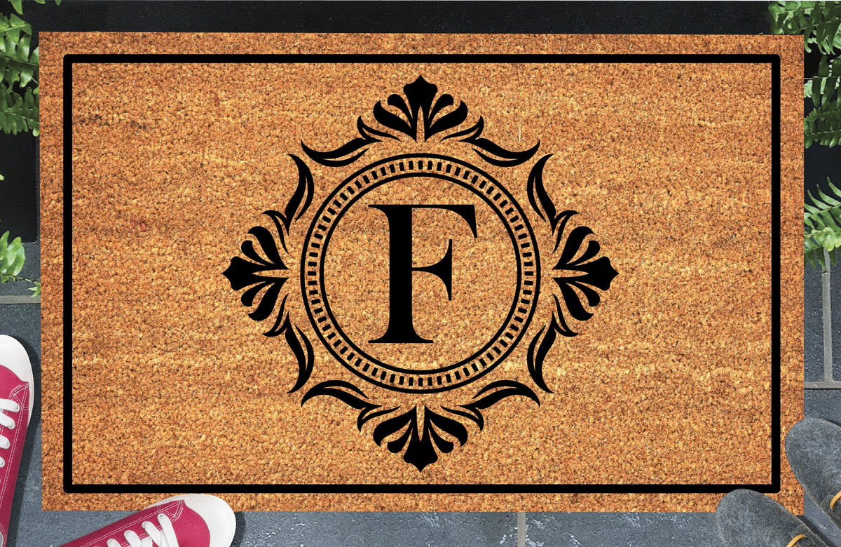 Monogram Letter F doormat in natural coir, showing the ornate serif letter F in a dotted circular frame with fleur-de-lis style flourishes and a black border, displayed on a doorstep. 