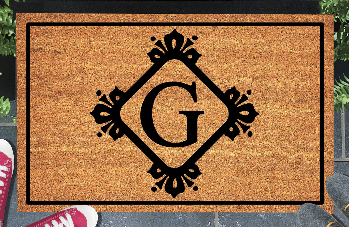 Monogram Letter G doormat in natural coir, featuring a serif G in a diamond frame with fleur-de-lis style flourishes, dot accents and a black border, displayed on a doorstep.  
