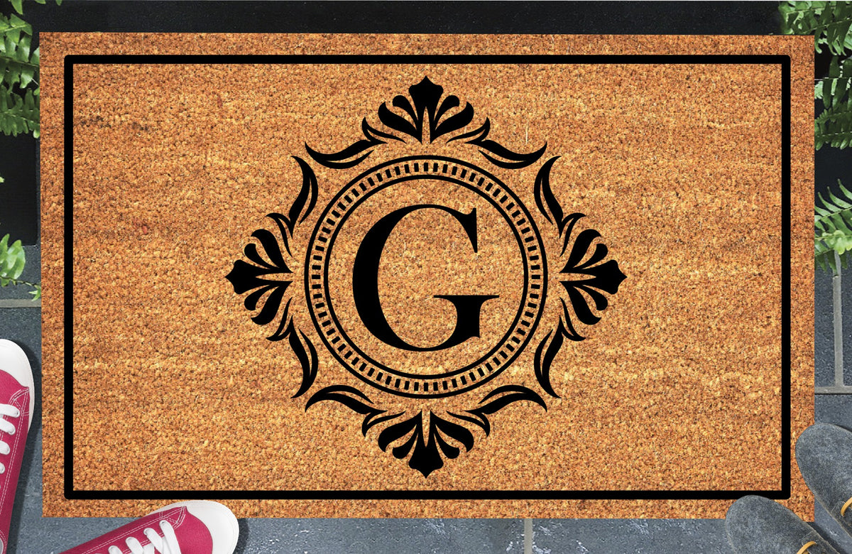 Monogram Letter G doormat in natural coir, showing the ornate serif letter G in a dotted circular frame with fleur-de-lis style flourishes and a black border, displayed on a doorstep.