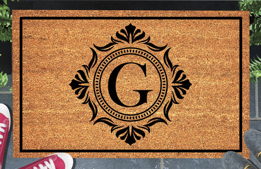 Monogram Letter G doormat in natural coir, showing the ornate serif letter G in a dotted circular frame with fleur-de-lis style flourishes and a black border, displayed on a doorstep.