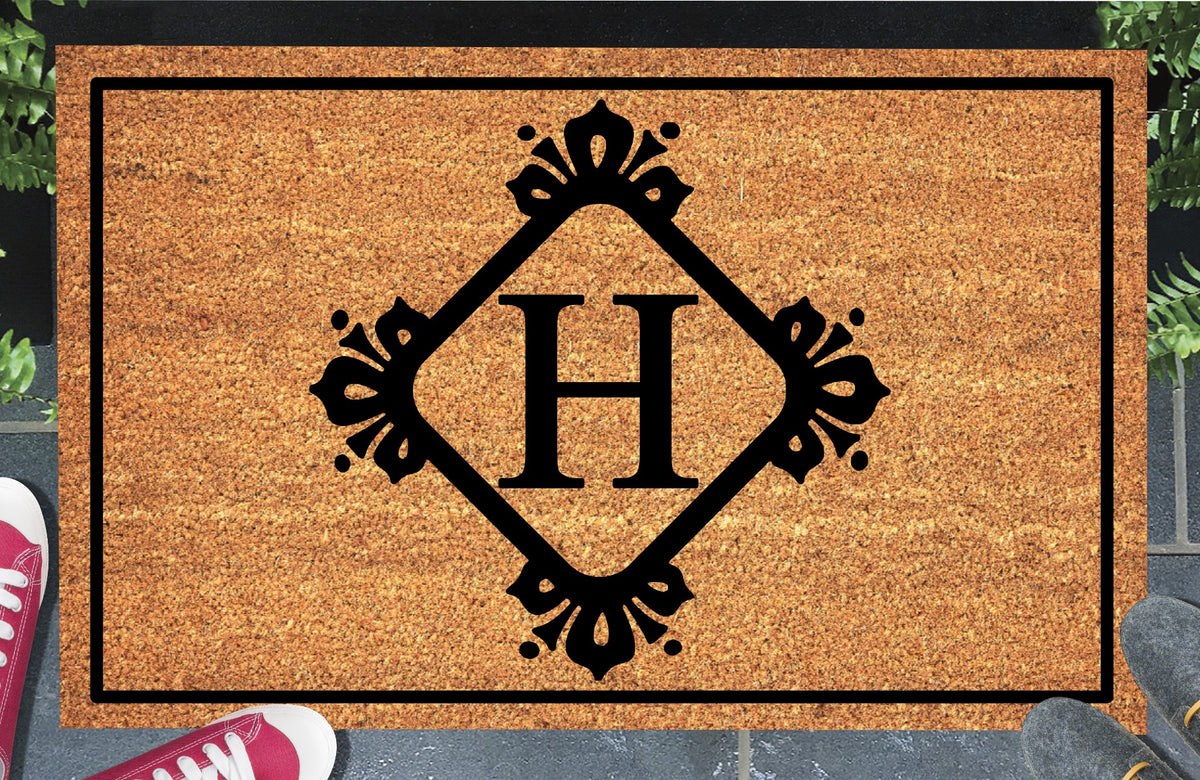Monogram Letter H doormat in natural coir, featuring a serif H in a diamond frame with fleur-de-lis style flourishes, dot accents and a black border, displayed on a doorstep. 
