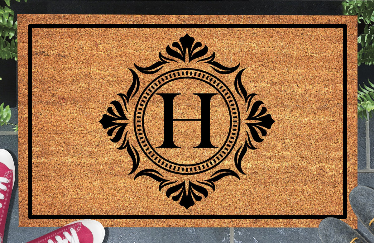 Monogram Letter H doormat in natural coir, showing the ornate serif letter H in a dotted circular frame with fleur-de-lis style flourishes and a black border, displayed on a doorstep.  