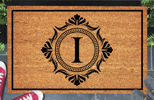 Monogram Letter I doormat in natural coir, showing the ornate serif letter I in a dotted circular frame with fleur-de-lis style flourishes and a black border, displayed on a doorstep. 