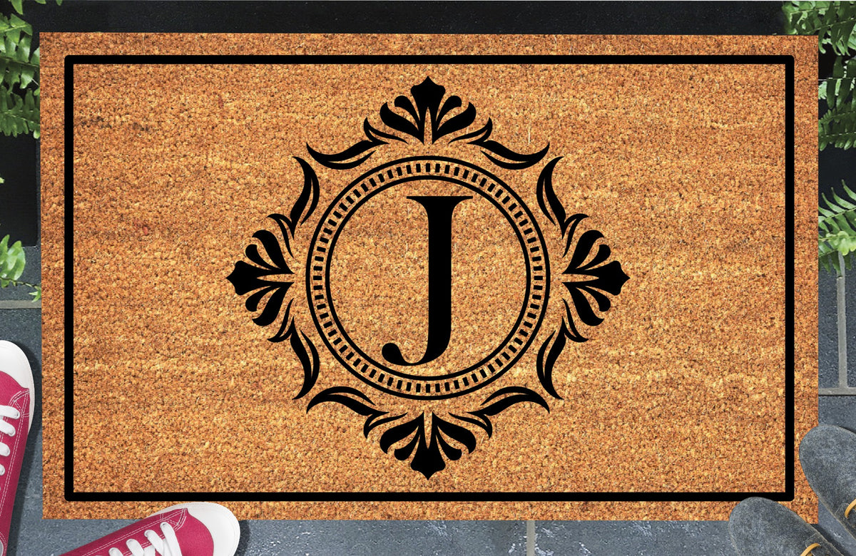 Monogram Letter J doormat in natural coir, showing the ornate serif letter J in a dotted circular frame with fleur-de-lis style flourishes and a black border, displayed on a doorstep. 
