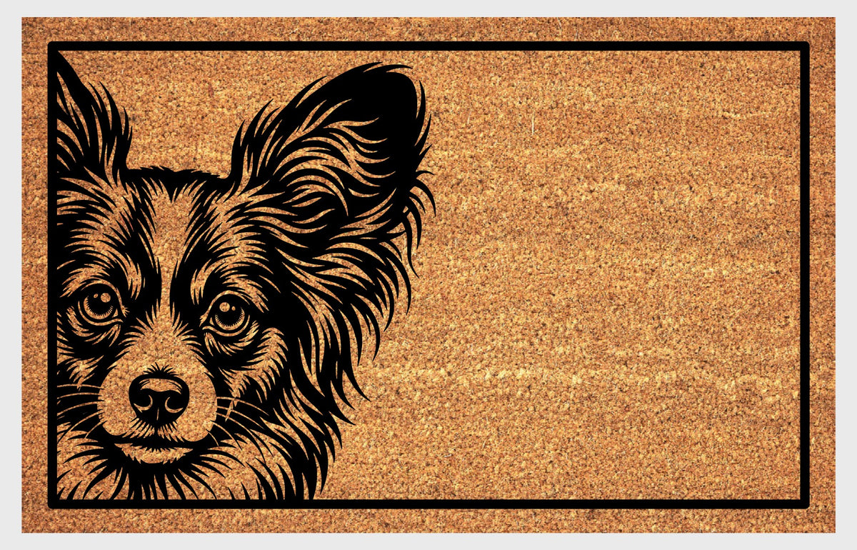 Papillon peeking from the left edge of a natural coir doormat, showing butterfly-shaped ears and bright eyes within a black rectangular border.