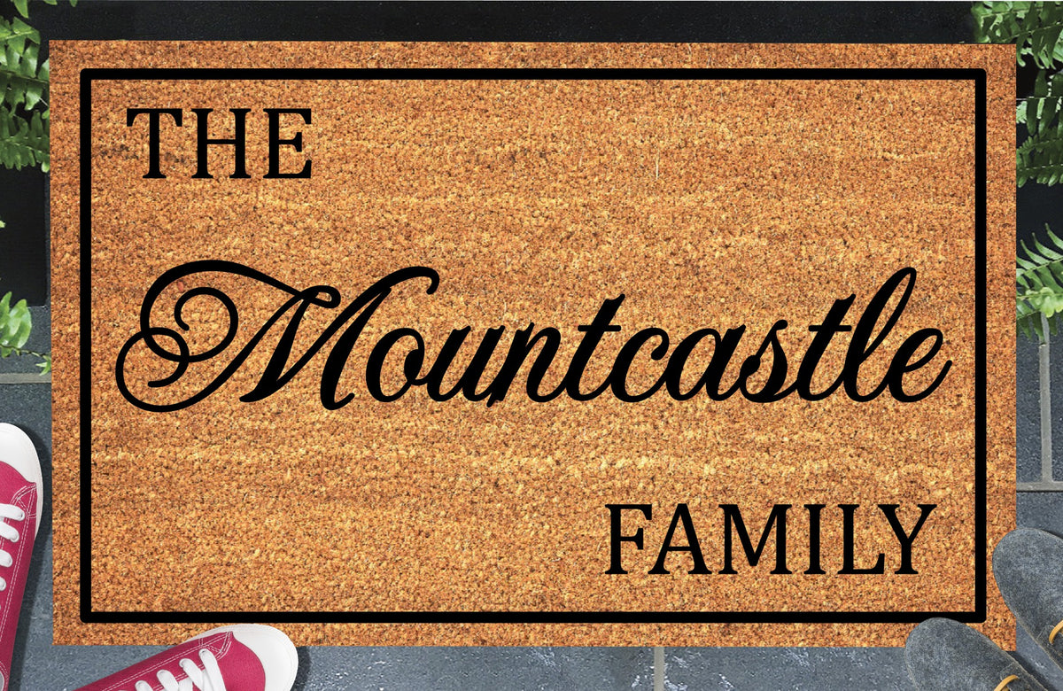 Personalized doormat with the family name in flowing black script inside a black border, displayed on a paved doorstep.