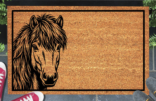 Shetland Pony peeking doormat in natural coir, featuring a black pony portrait peeking from the left edge with small upright ears, expressive eyes, a heavy forelock and a shaggy mane inside a black rectangular border, displayed on a doorstep.