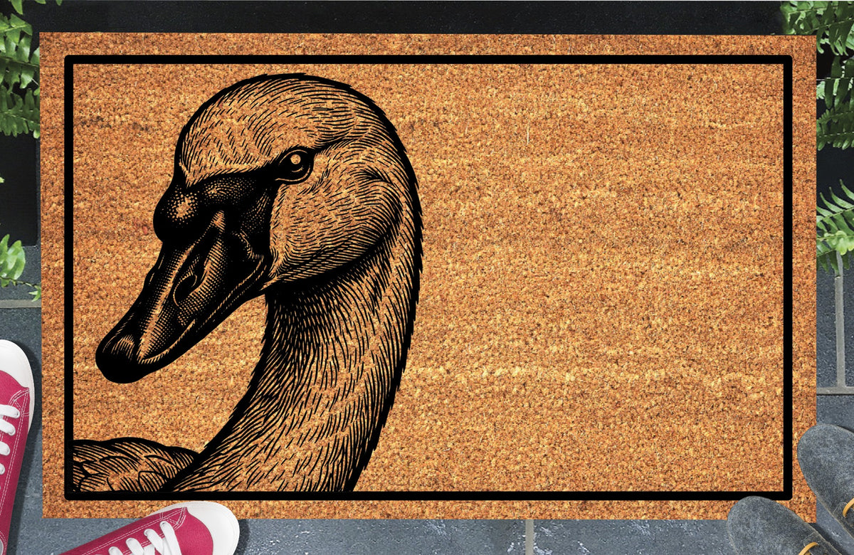 Trumpeter Swan peeking from the left edge of a natural coir doormat, with a curved neck, detailed feathers and black bill angled downwards, within a black border at a doorstep.